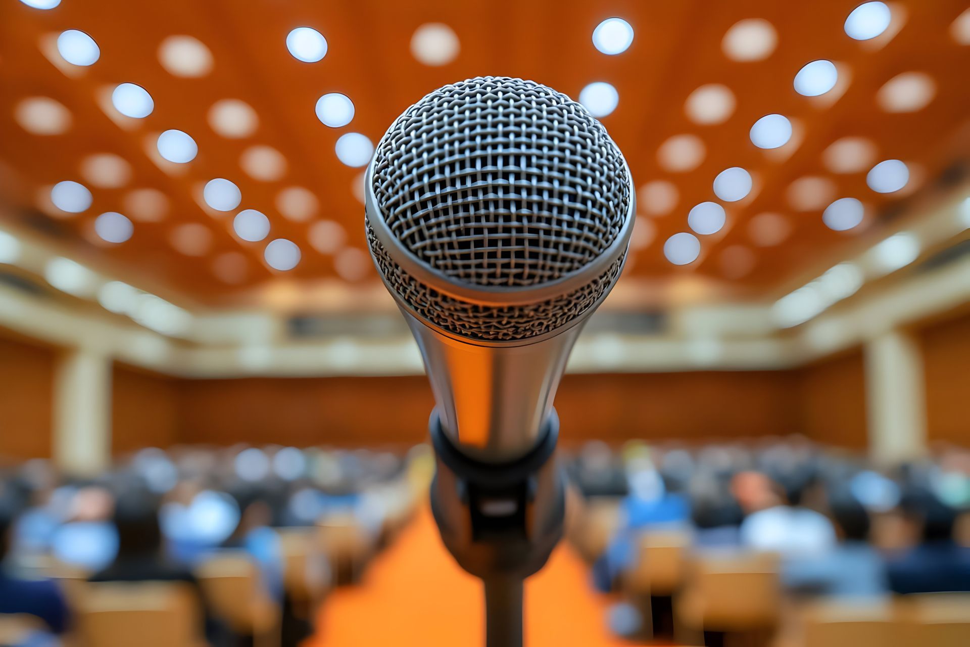 Un microphone placé au premier plan, face à un public assis dans un auditorium flou aux tons chauds.