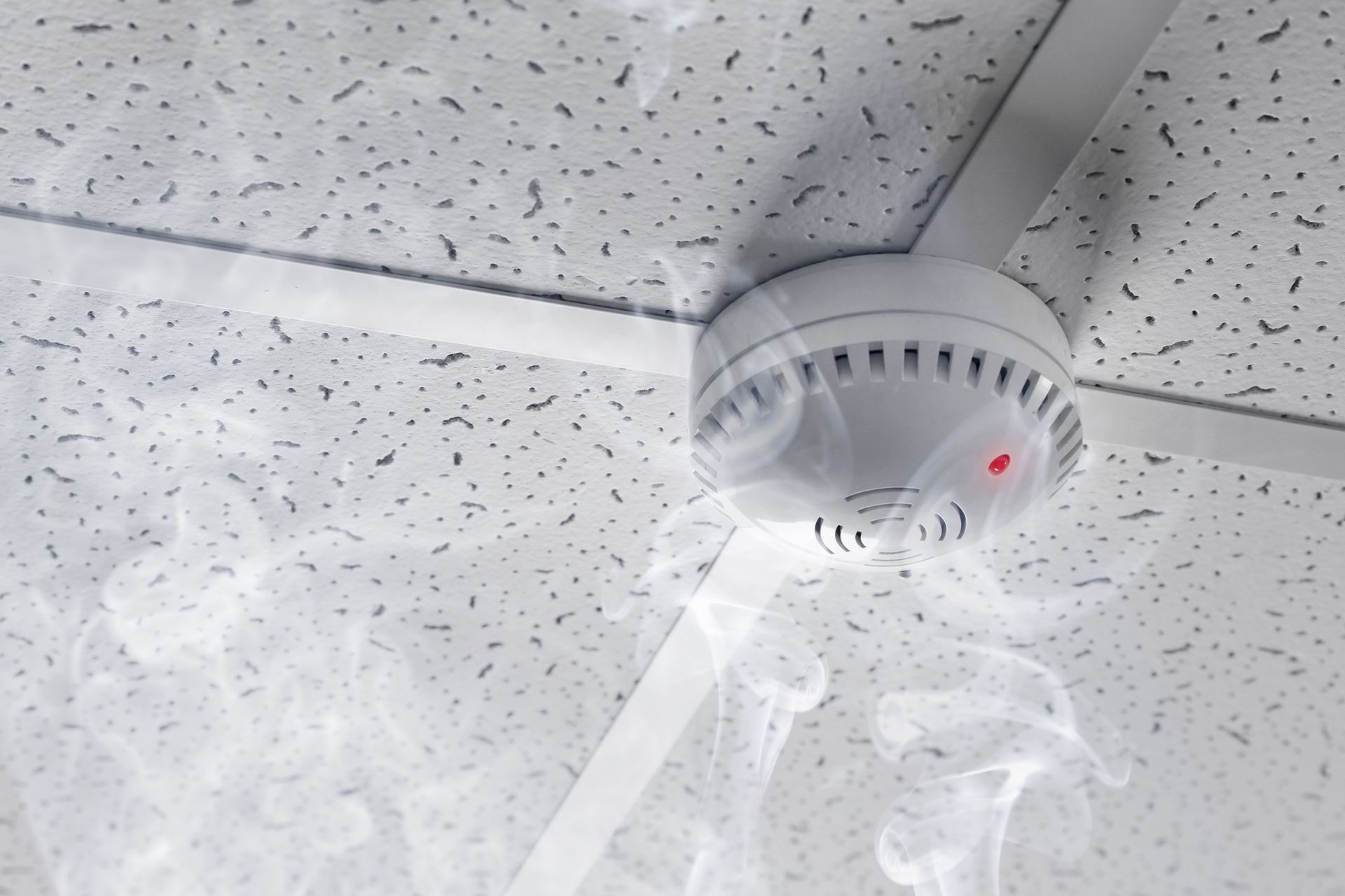 Un détecteur de fumée fixé sur un faux plafond blanc, d'où s'échappent des volutes de fumée et une lumière rouge.