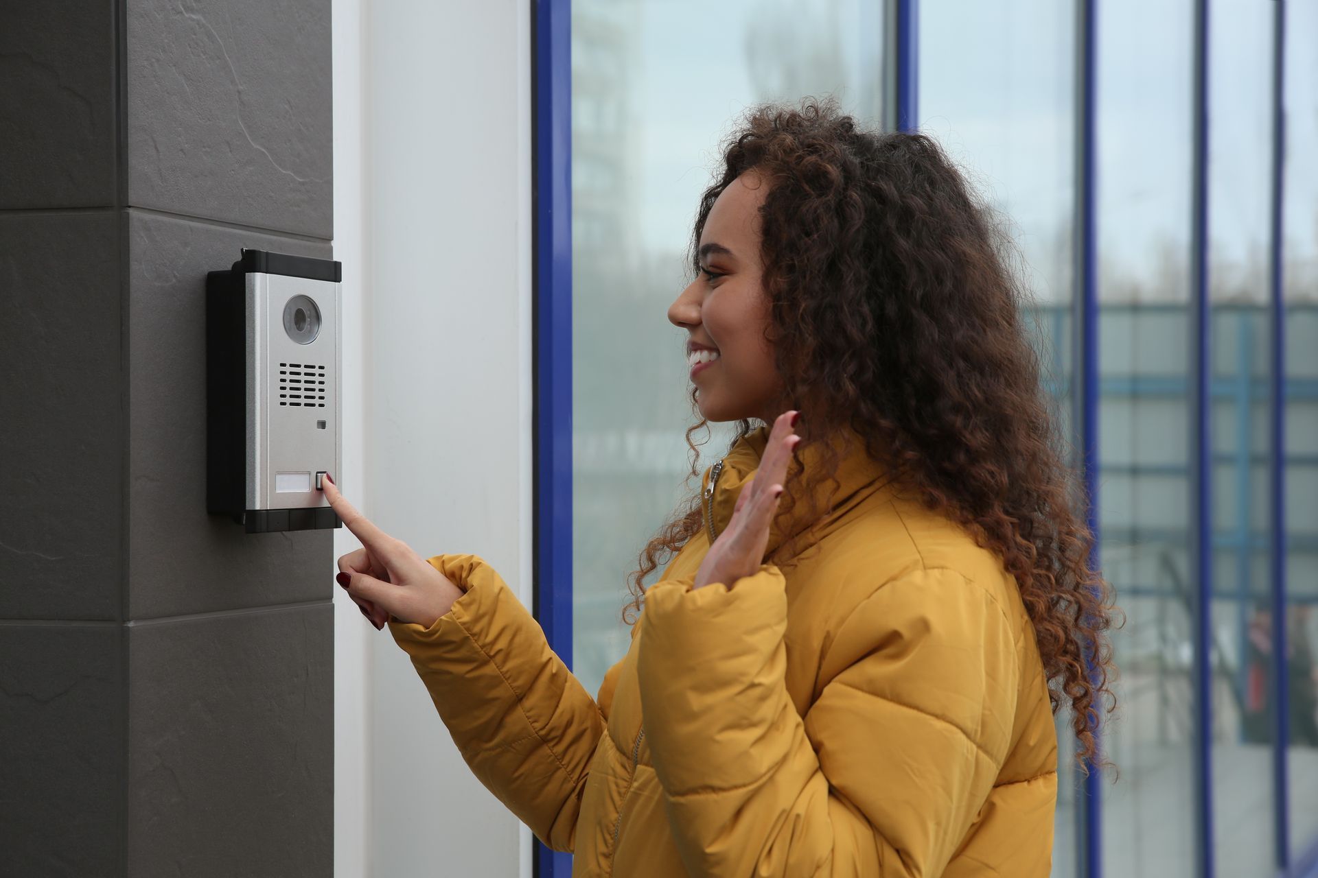 Une personne appuie sur un bouton d'un interphone mural, souriant et faisant un signe de la main en direction de l'appareil.