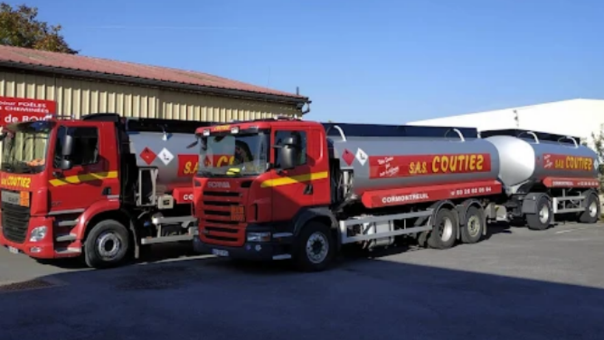 Deux camions-citernes Scania rouges garés côte à côte sur un parking goudronné devant un bâtiment industriel beige, sous un ciel bleu.