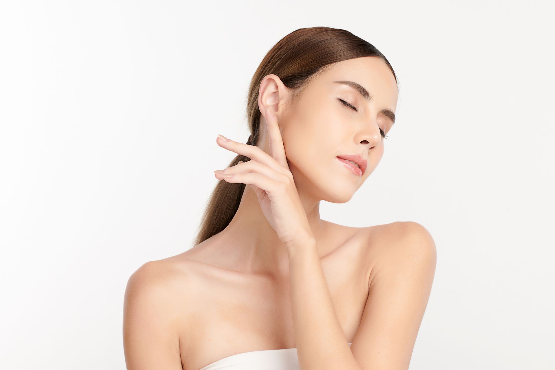 Femme aux longs cheveux bruns, les yeux fermés, posant sur un fond blanc et touchant sa nuque.