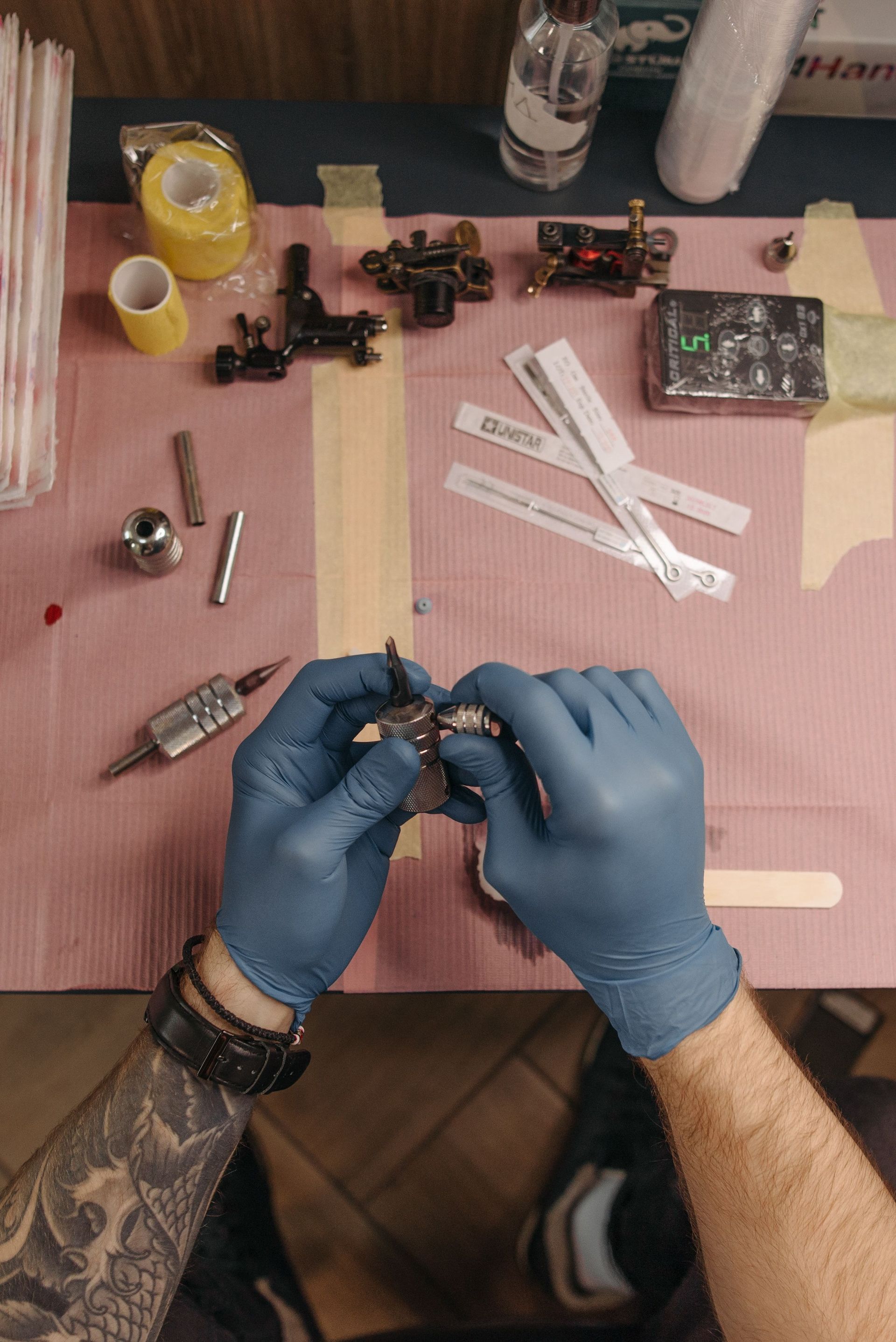Tatoueur portant des gants bleus en train d'assembler une machine à tatouer au-dessus d'un poste de travail recouvert de rose, avec des outils et des godets d'encre.