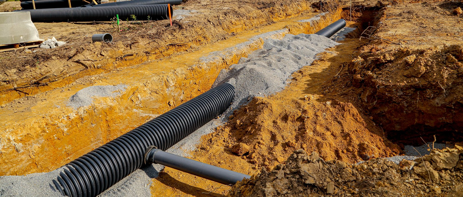 Une pelleteuse travaille sur un chantier boueux où de gros tuyaux de drainage noirs sont posés dans des tranchées sous un ciel bleu.