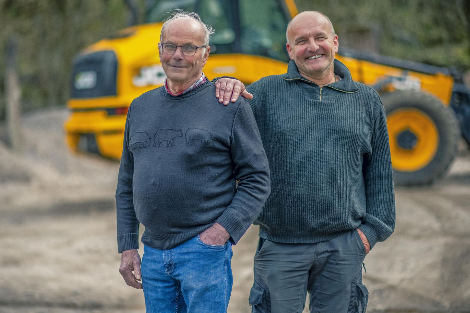 Deux hommes debout à l’extérieur devant un engin de chantier jaune, l’un passant un bras autour de l’autre.