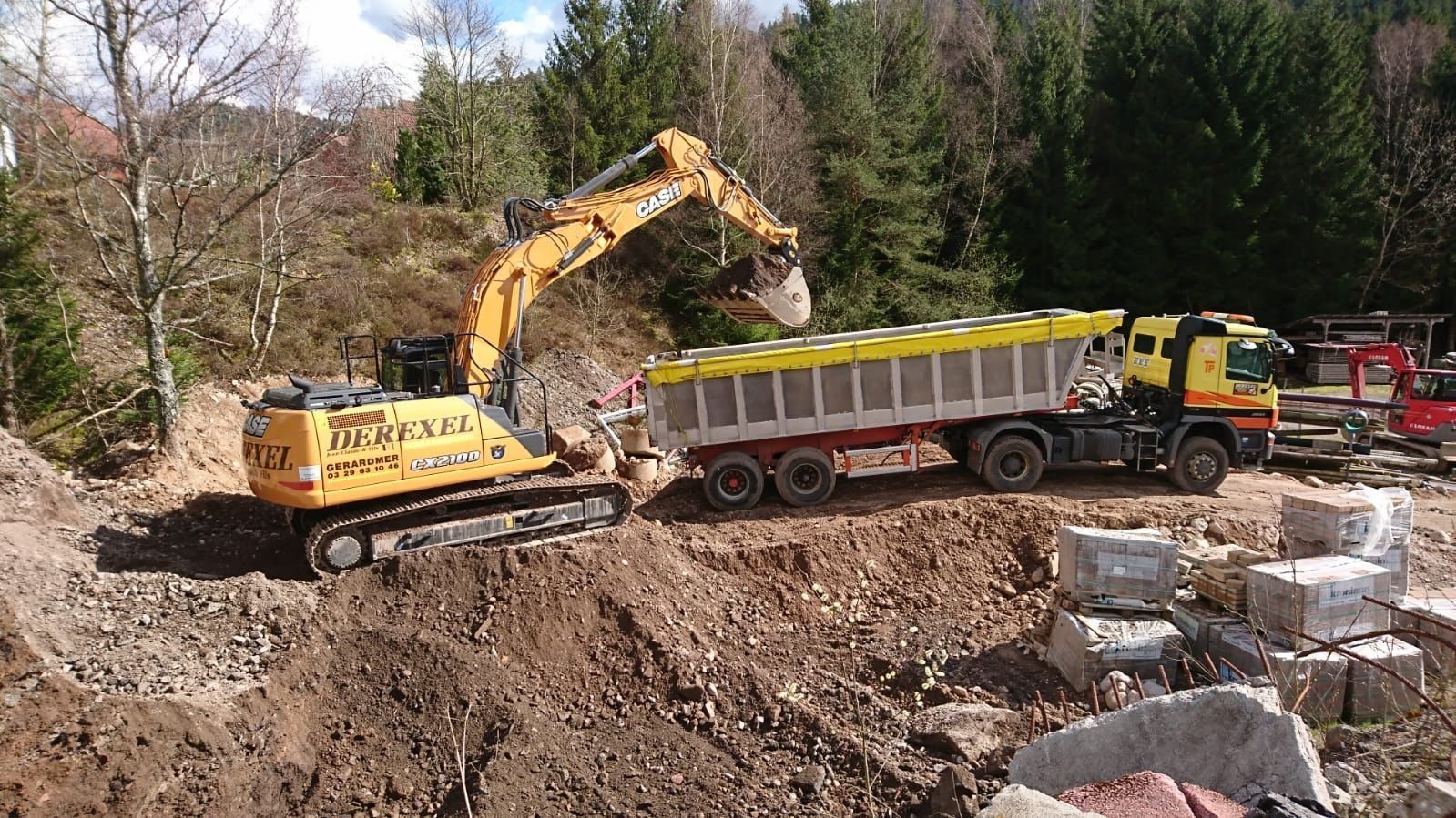 Une excavatrice charge de la terre dans un camion-benne sur un chantier, avec des arbres et des rochers à côté.