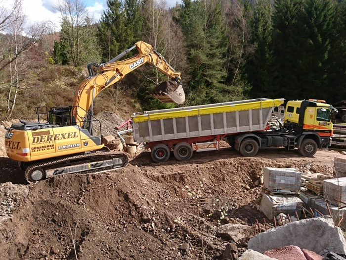 Une pelleteuse chargeant de la terre dans un camion-benne sur un chantier entouré d’arbres.