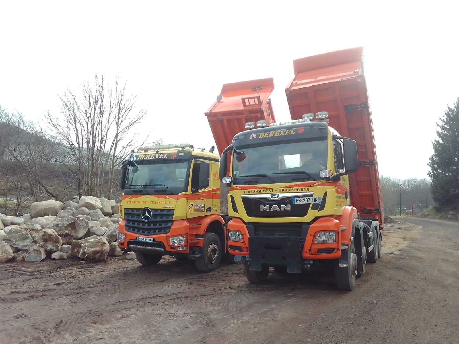 Deux camions-bennes orange circulent sur un chemin de terre, leurs bennes levées, tandis que du gravier ou des pierres se trouvent à côté.