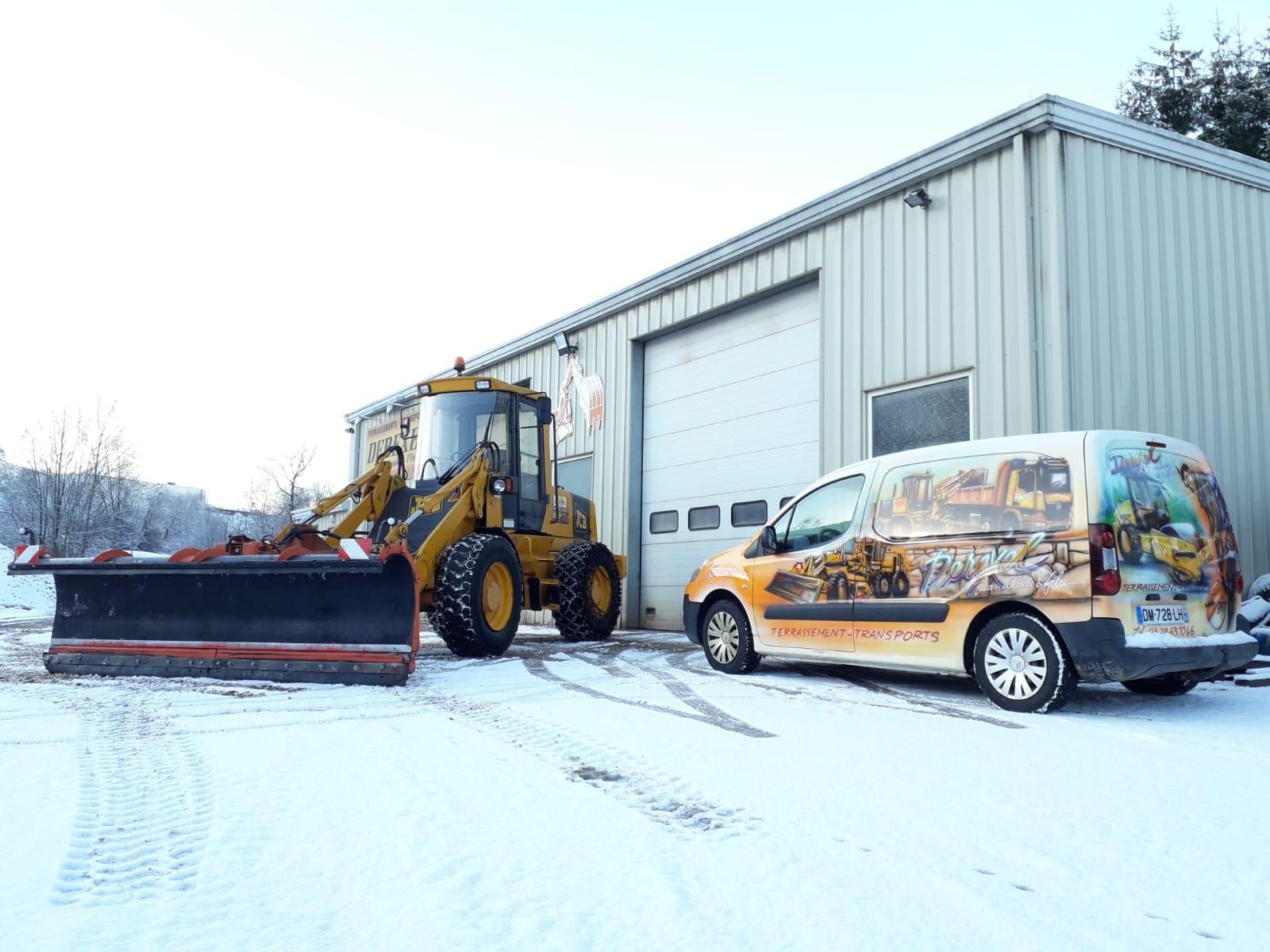 Un chasse-neige jaune est garé à côté d’une camionnette, devant un entrepôt, sur un terrain enneigé.