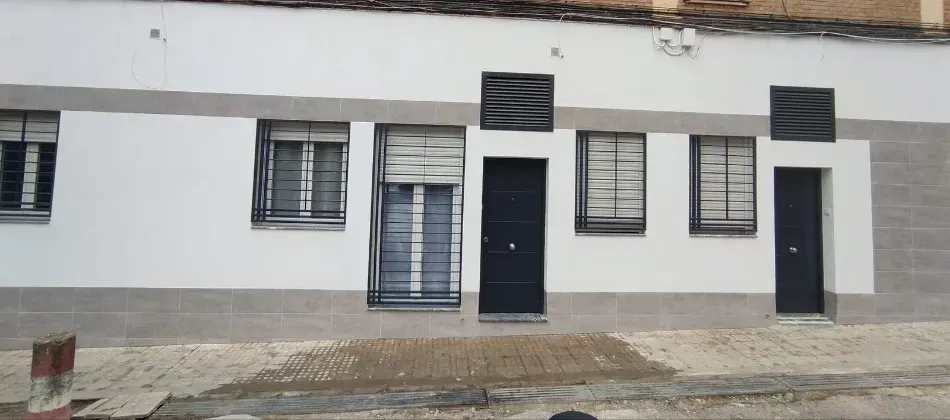 Fachada blanca de un edificio con ventanas enrejadas y dos puertas oscuras en una calle pavimentada.