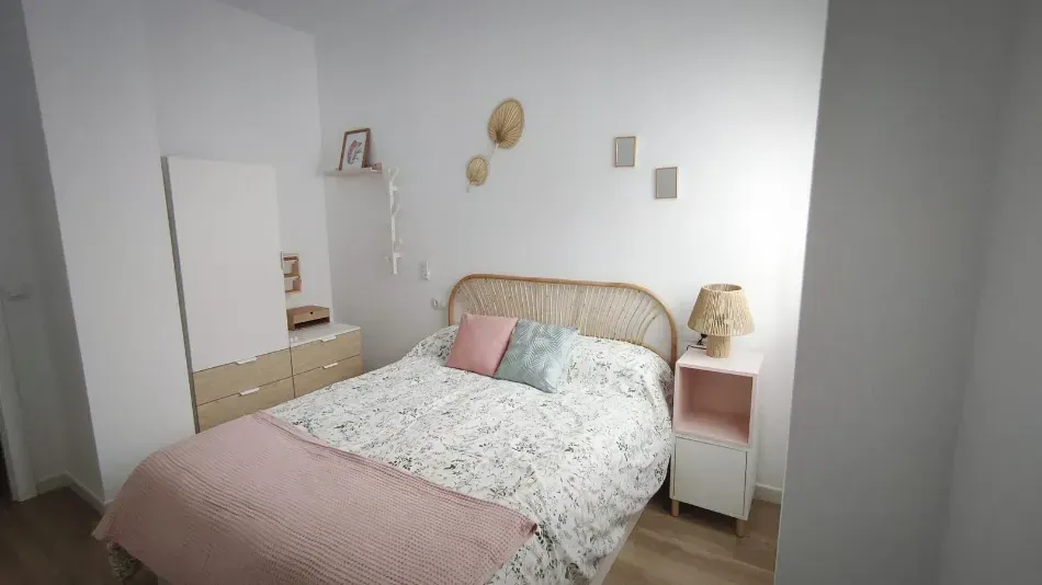 Habitación luminosa con cama blanca, ropa de cama rosa, mesitas de noche y armario en la esquina.