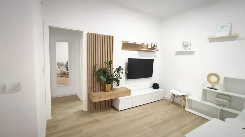 Sala de estar luminosa y minimalista con televisor, pared decorativa revestida de listones de madera, estantes blancos y suelo de madera clara.
