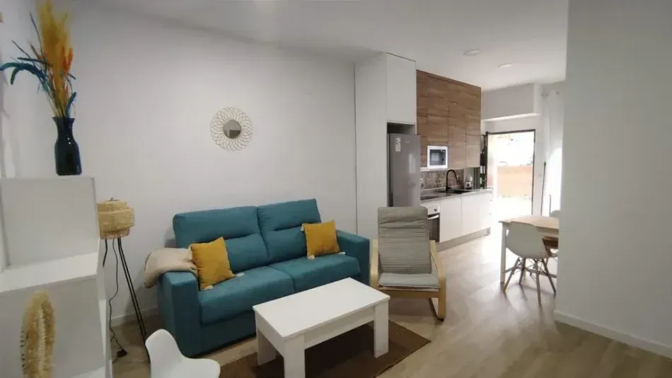 Sala de estar moderna con sofá color turquesa, mesa de centro blanca, sillón y área de cocina abierta.