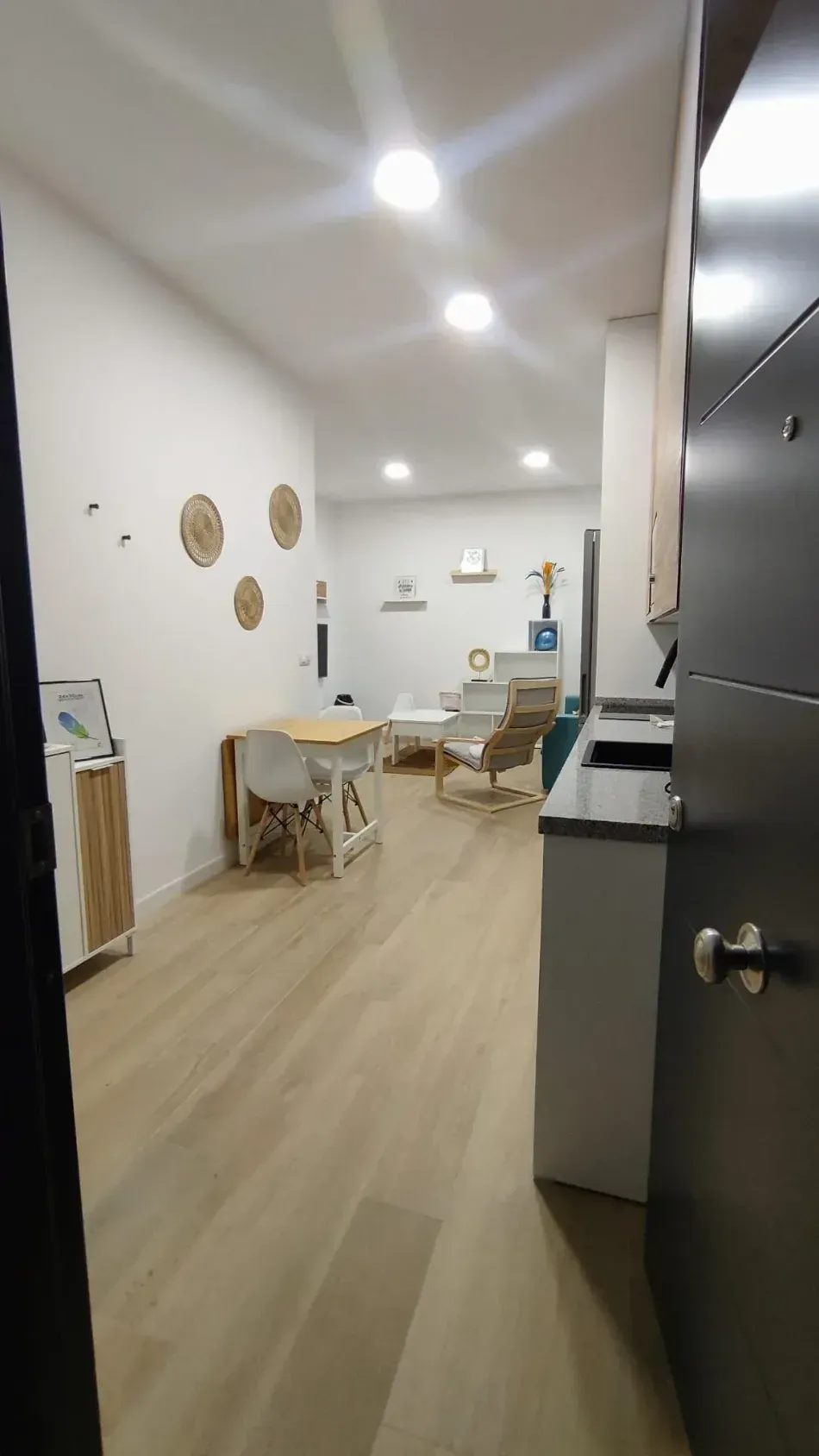 Comedor luminoso y minimalista con sillas de madera, paredes blancas y luces empotradas en el techo, visto desde una puerta.