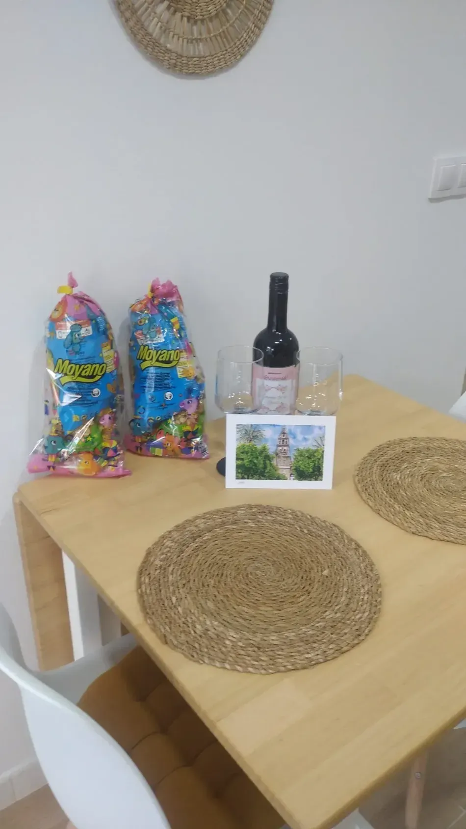 Mesa de comedor con manteles individuales tejidos, coloridas bolsas de regalo, una botella de vino y una pequeña tarjeta con una foto encima.