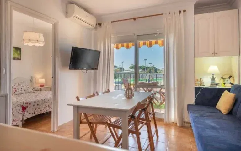 Luminoso comedor en el apartamento con mesa, sofá azul, televisor de pared y vista al balcón a través de las puertas abiertas.