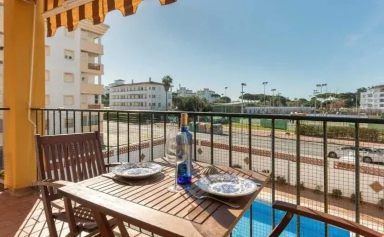 Balcón con mesa de madera, sillas y mesa para desayunar con vistas a una cancha de tenis y edificios.