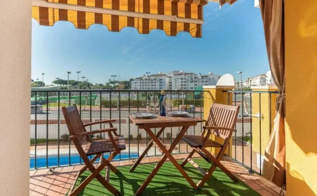 Terraza soleada con mesa y sillas de madera, toldo a rayas y vistas a la ciudad más allá de la barandilla.