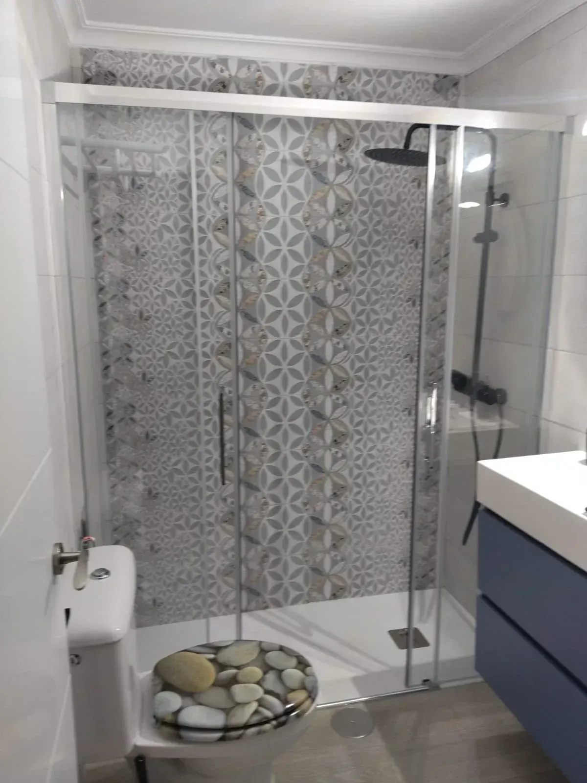 Baño moderno con cabina de ducha de cristal, azulejos estampados, inodoro y lavabo con mueble.