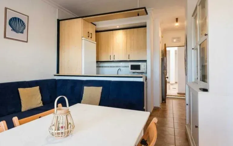 Cocina luminosa y rincón de comedor con bancos azules, mesa blanca y armarios de madera clara en un apartamento tipo pasillo.