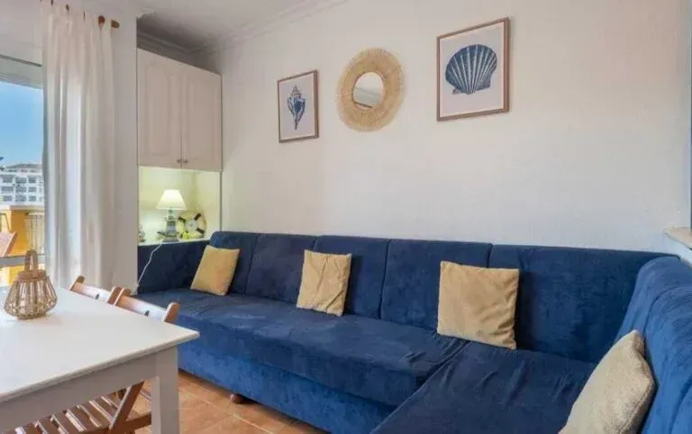 Sala de estar luminosa con un largo sofá azul, una mesa de centro blanca y cuadros con motivos costeros en las paredes.
