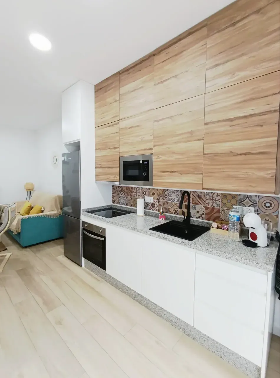 Cocina moderna con gabinetes blancos, pared revestida de madera, encimera negra y un sofá color turquesa al fondo.