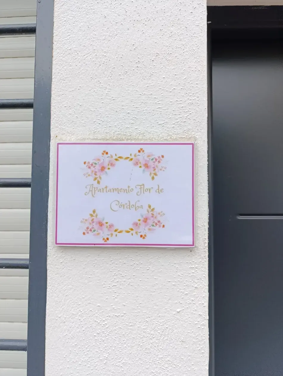 Letrero floral con borde rosa y texto cursivo dorado montado sobre un pilar blanco en el exterior.