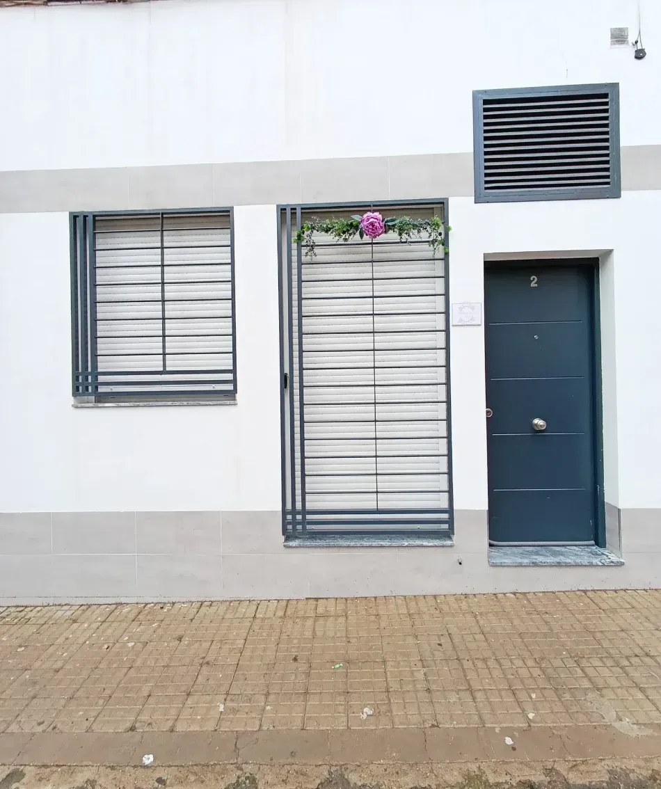 Fachada de un edificio moderno de color blanco con dos ventanas enrejadas, una puerta azul y una rejilla de ventilación en la parte superior, vista desde la acera.