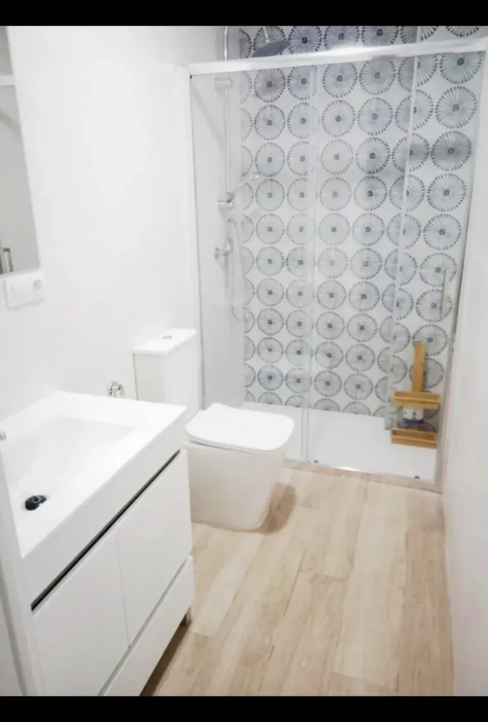 Baño pequeño y moderno con lavabo blanco, inodoro, suelo de madera y cortina de ducha estampada.