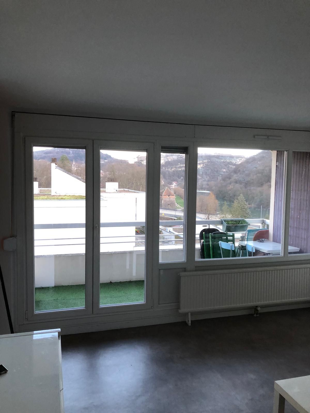 Grandes fenêtres dans une pièce proposant une vue sur les bâtiments et les arbres. Boiseries, radiateur et sol.