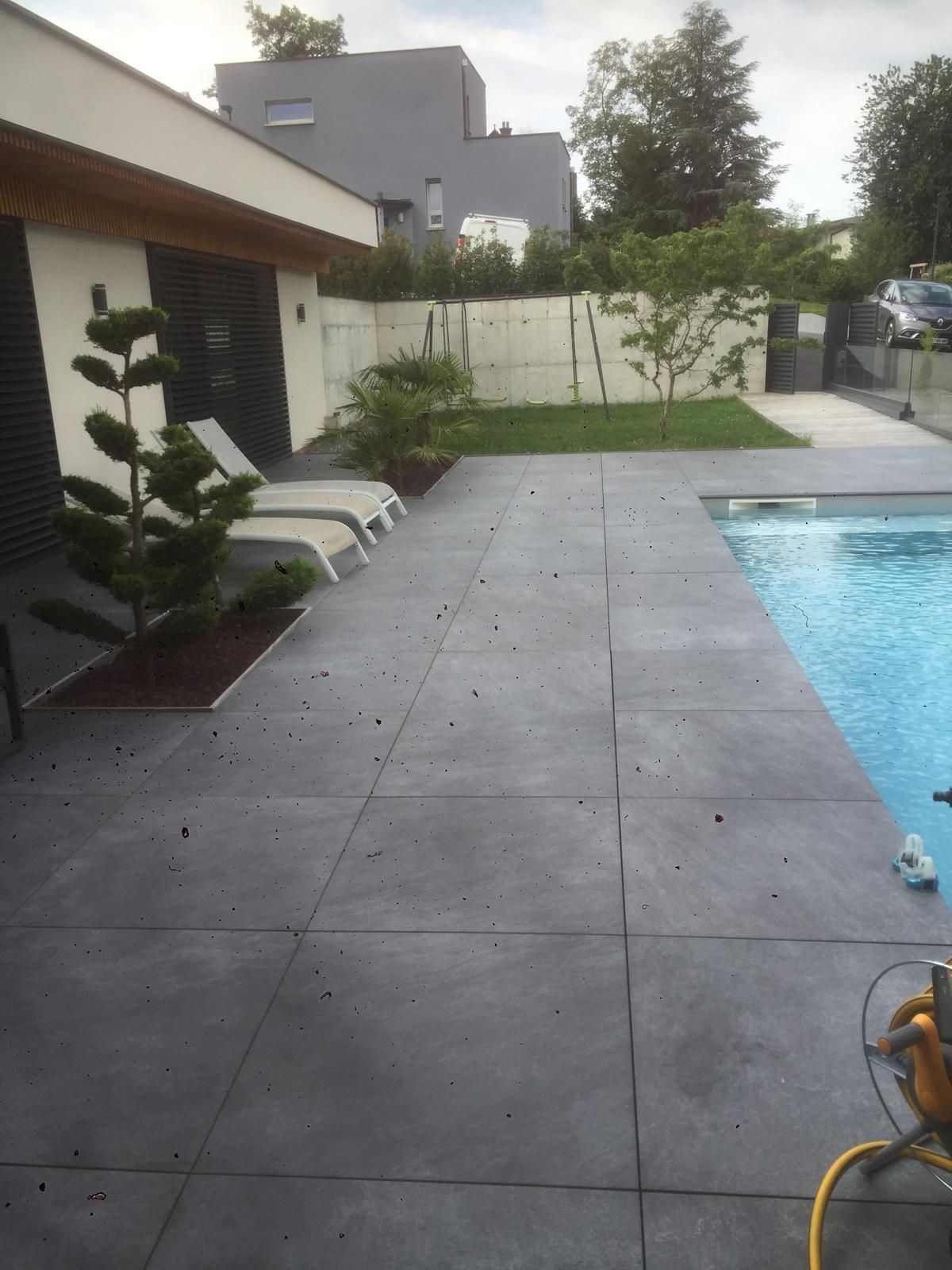 Terrasse carrelée donnant sur une piscine et une pelouse. Arbres, chaises longues et bâtiment visibles.