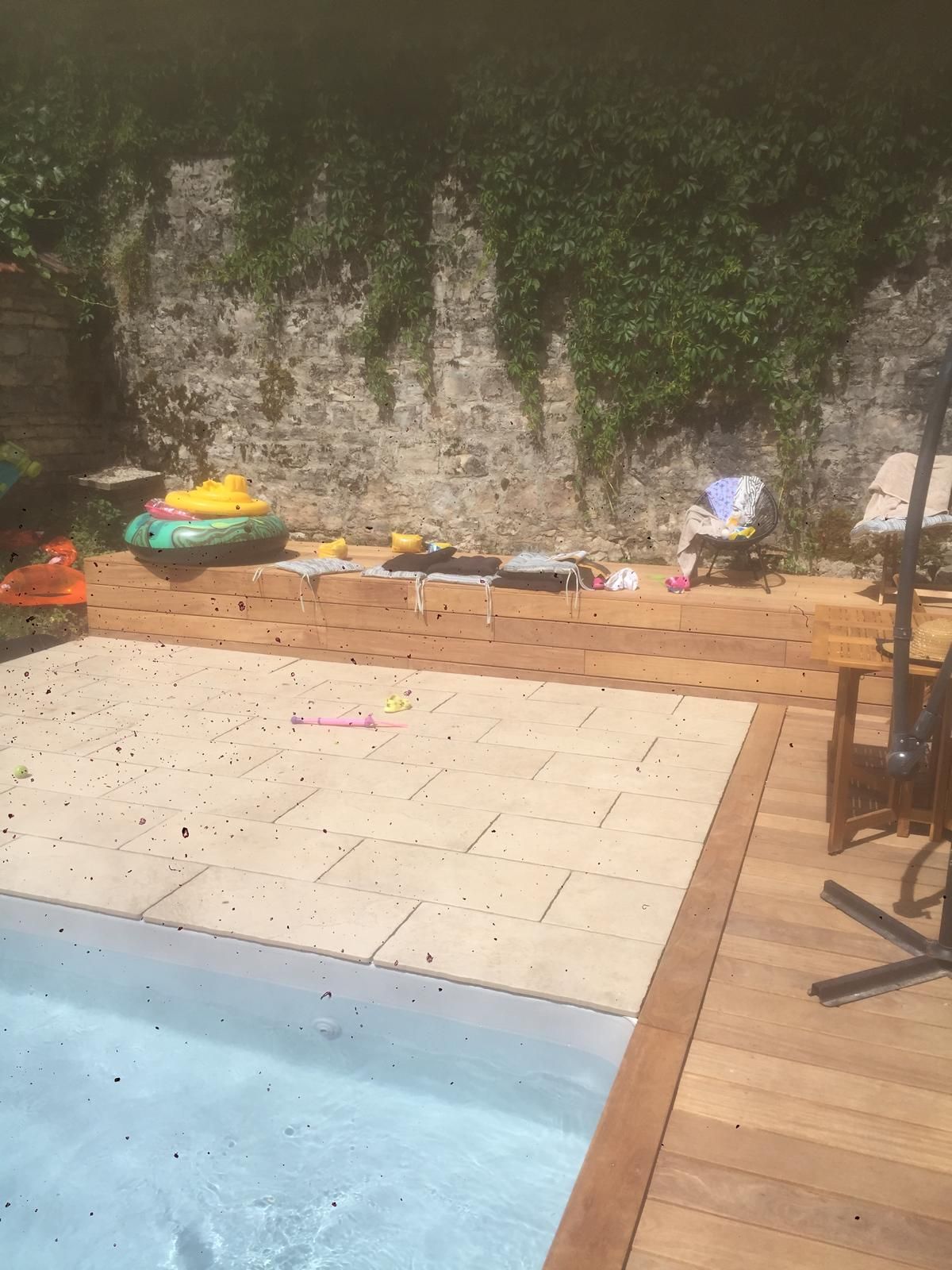 Paysage de jardin avec piscine, terrasse en pierre, terrasse en bois et mur de briques recouvert de lierre.