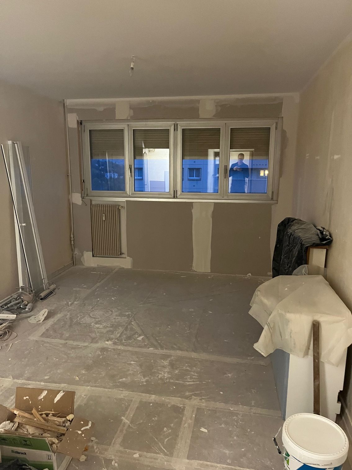 Pièce en rénovation avec cloisons sèches, fenêtre, débris au sol et seau de peinture.