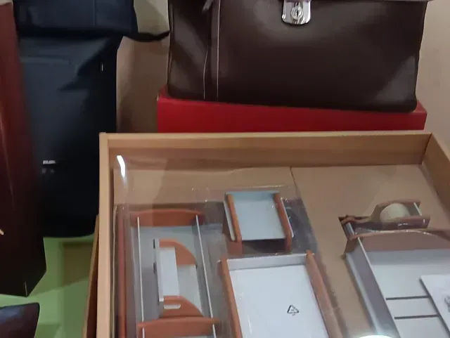 Una exhibición de artículos de oficina nuevos. Un maletín de cuero marrón reposa sobre una caja roja. Debajo, una caja de madera con tapa transparente contiene organizadores de oficina.