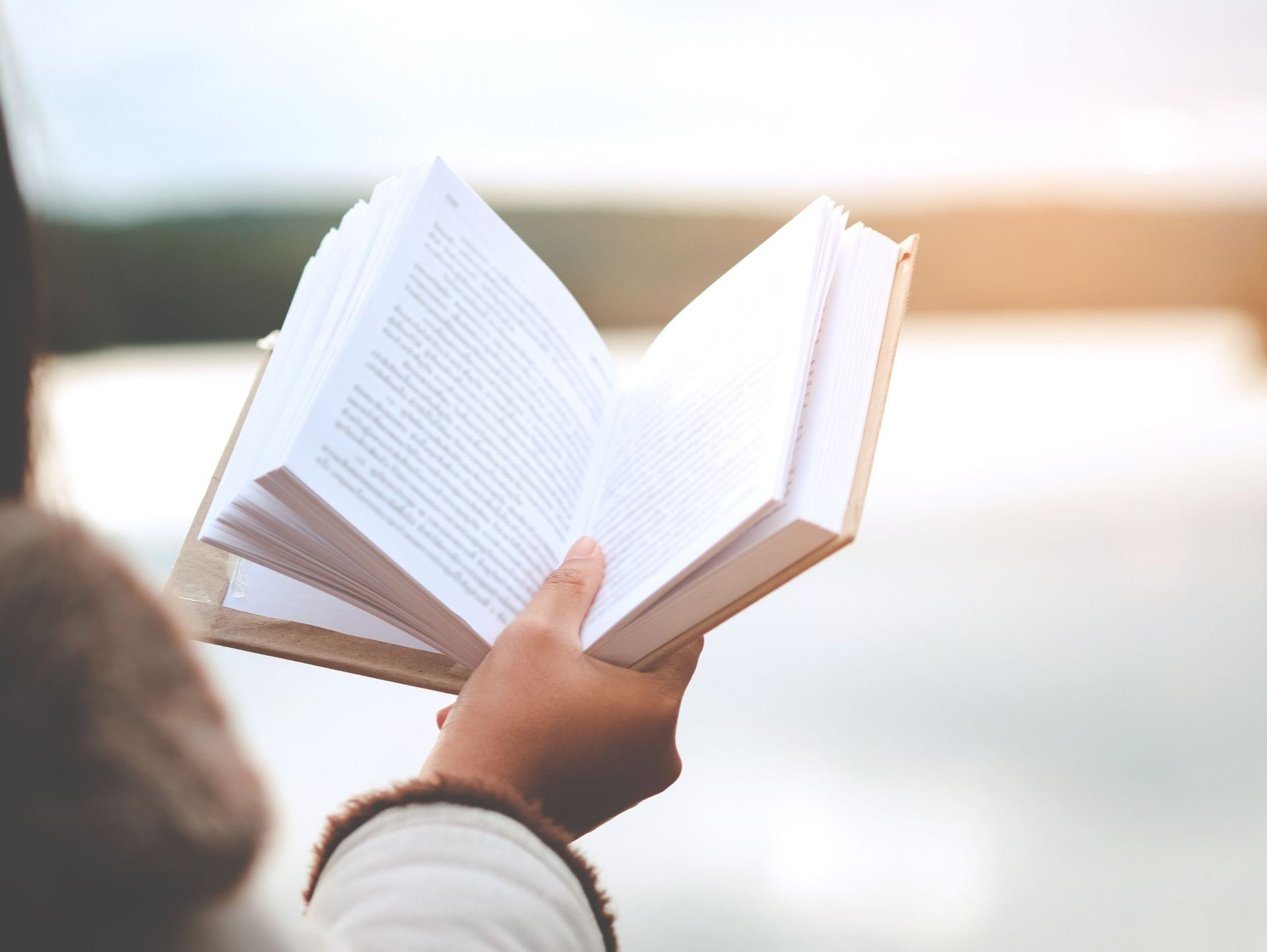 Persona sosteniendo un libro abierto cuyas páginas se ven contra un fondo exterior borroso. Luz del sol a la derecha.