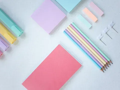 Útiles de oficina de colores pastel: notas adhesivas, resaltadores, lápices y chinchetas dispuestos sobre una superficie blanca.