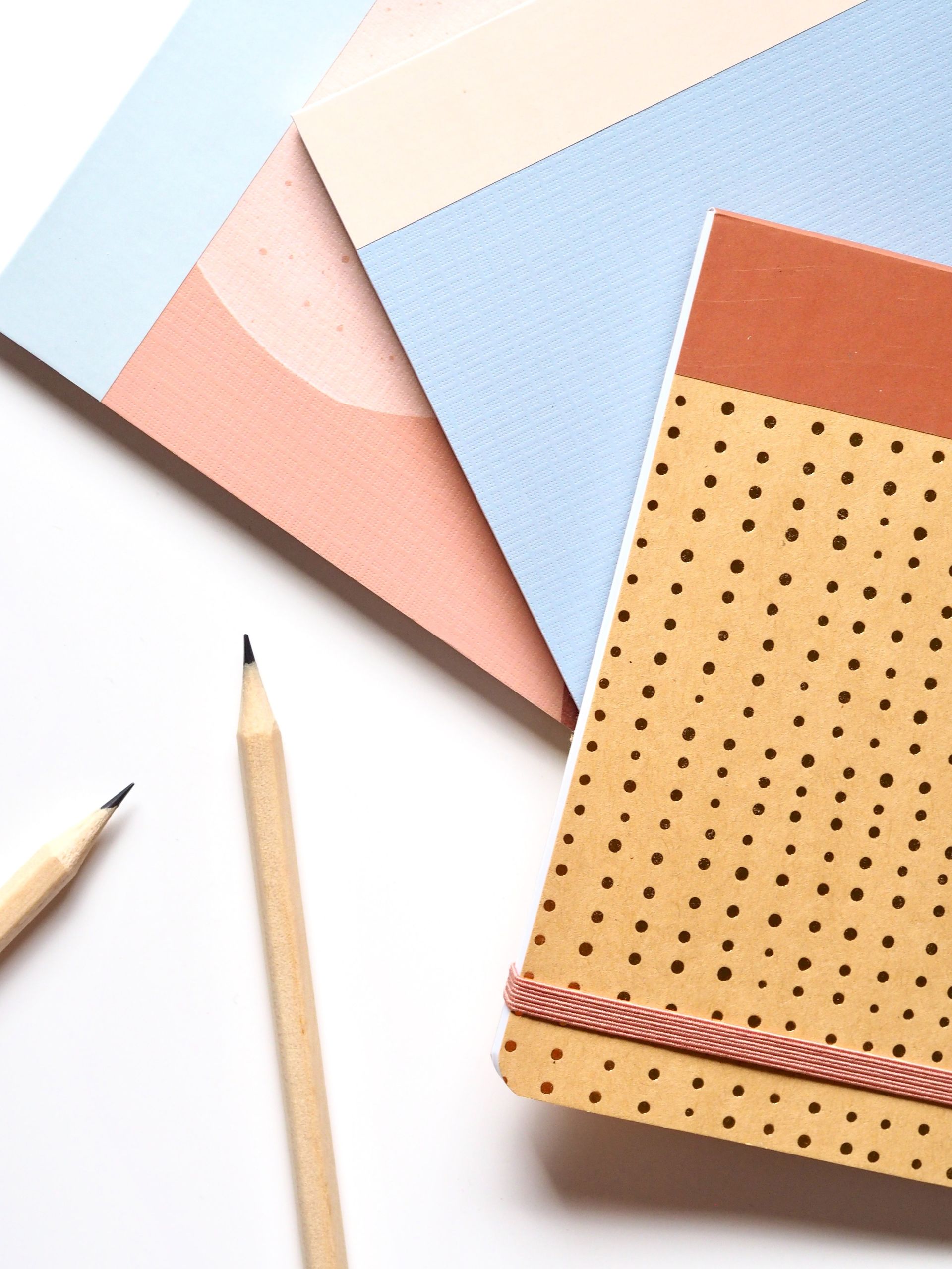 Cuadernos y lápices sobre una superficie blanca. Hay papeles geométricos de colores pastel y un cuaderno tostado con lunares.