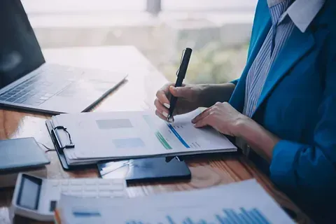 Mujer con chaqueta azul escribiendo en papeles en un escritorio con computadora portátil, calculadora y teléfono.