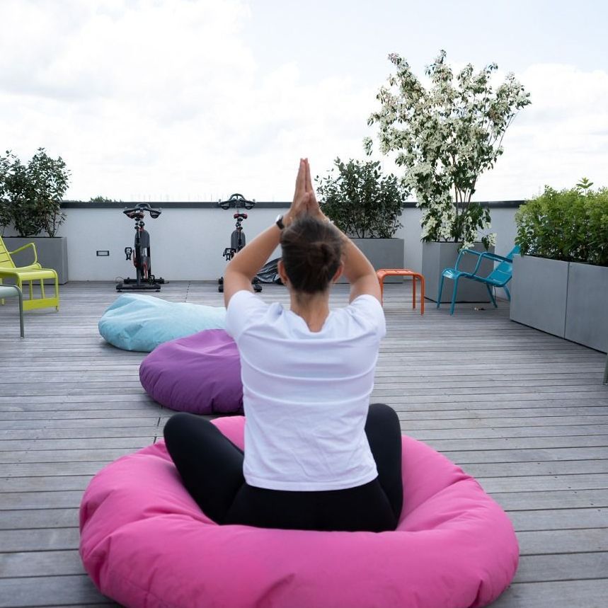 Person in weißem Hemd und schwarzen Leggings macht im Yoga auf einem rosa Sitzsack. Sie blickt weg und hat die Hände gefaltet