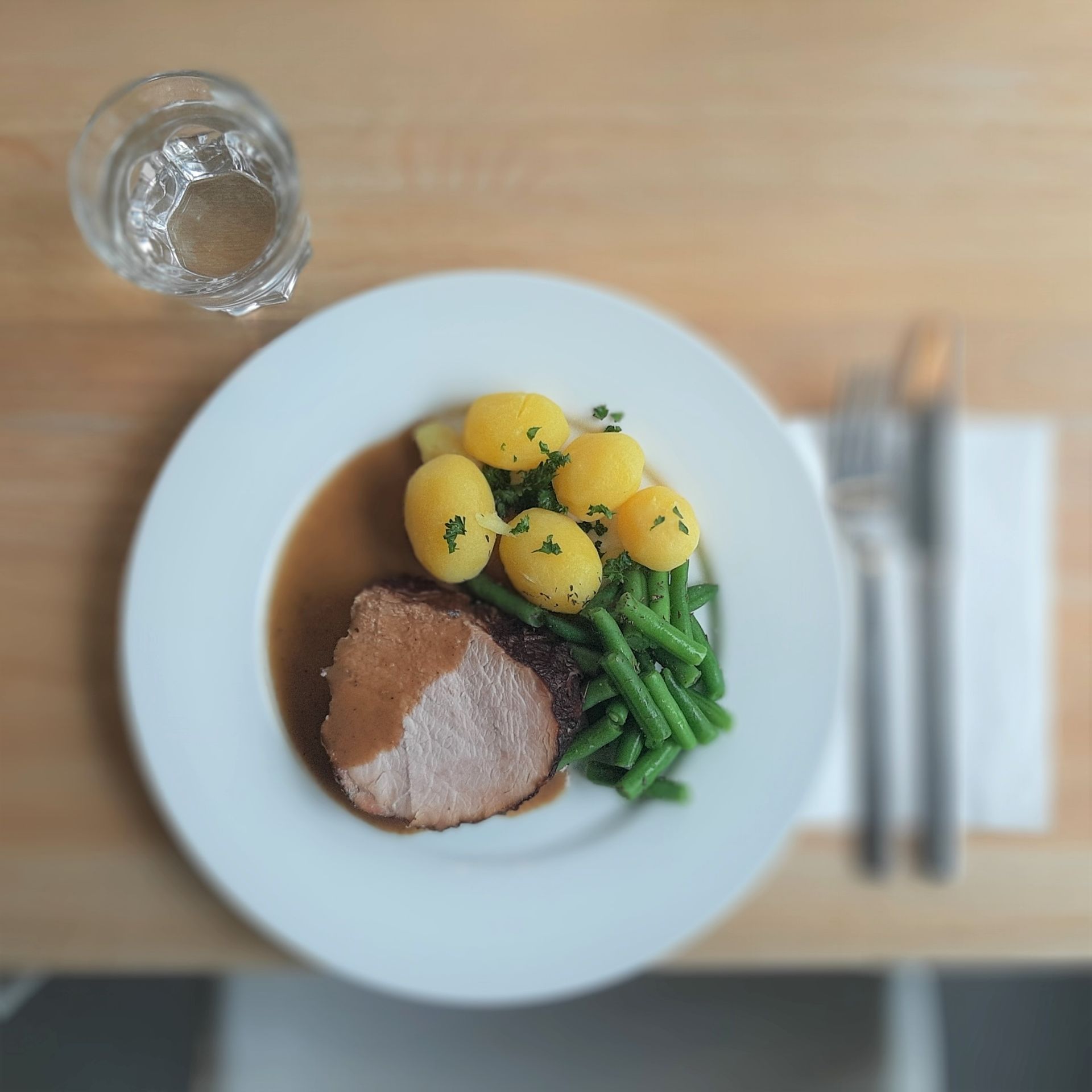 Teller mit Braten mit Soße, Salzkartoffeln und grünen Bohnen, daneben ein Glas Wasser und Besteck auf einem Holztisch.
