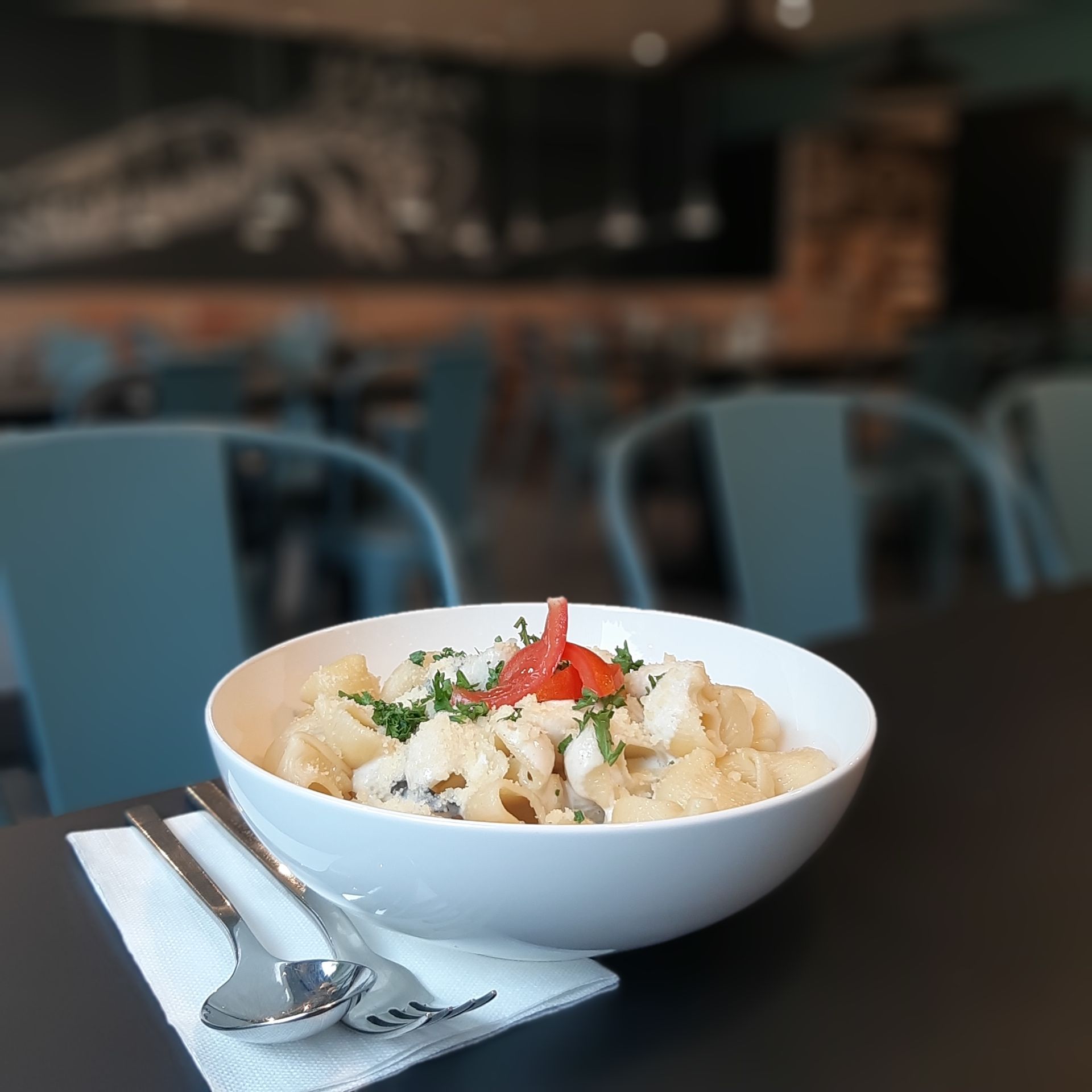 Eine weiße Schüssel Pasta mit Tomatengarnitur, Besteck und einer Serviette auf einem schwarzen Tisch in einem Restaurant.