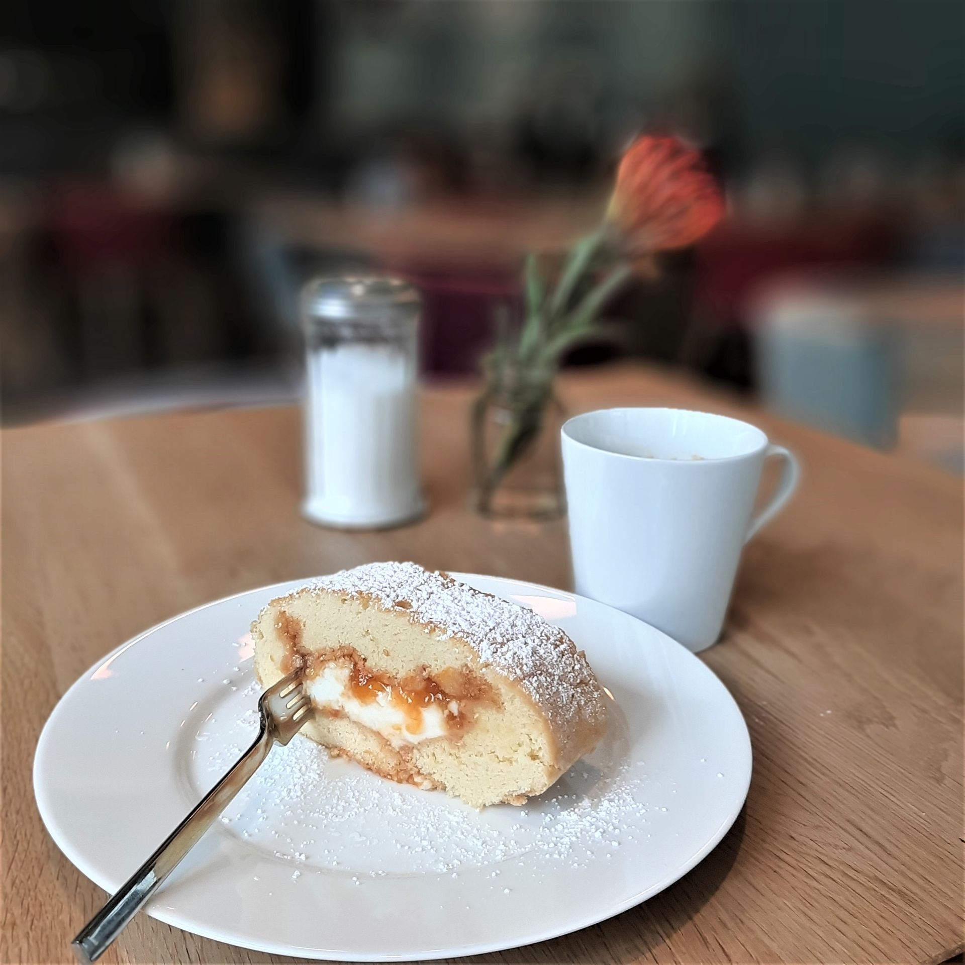 Eine Biskuitrolle mit Puderzucker auf einem weißen Teller, neben einer Tasse und einem Zuckerspender auf einem Holztisch.