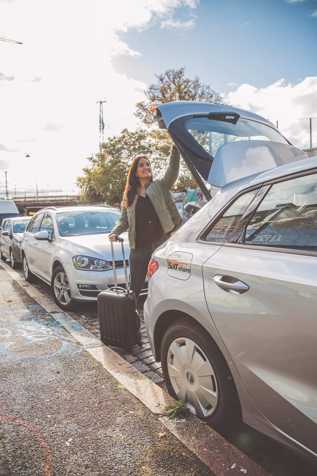 Woman loading luggage into a silver car trunk on a sunny day. She's smiling, next to a parked line of cars.
