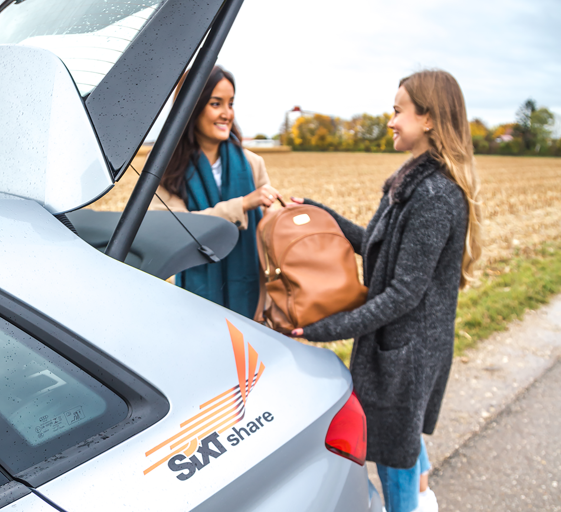 Two women loading a backpack into the trunk of a Sixt share car. They are smiling in a rural setting.