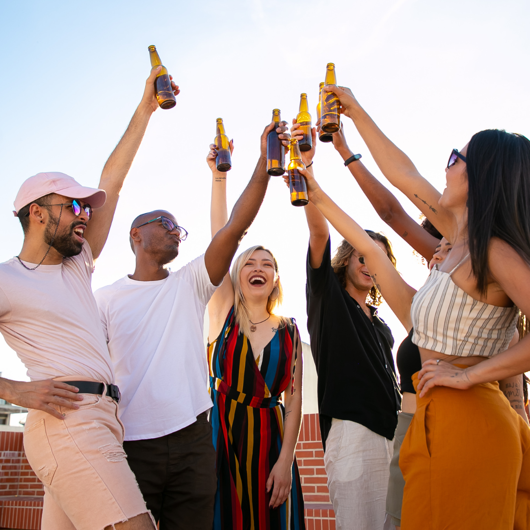 A diverse group cheers with beer bottles held high on a rooftop on a sunny day.