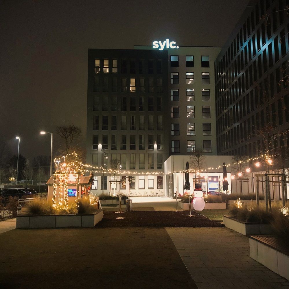Nachtszene vor einem Gebäude mit „Sylc“-Beschilderung. Weihnachtslichter und ein Schneemann schmücken den Hof.