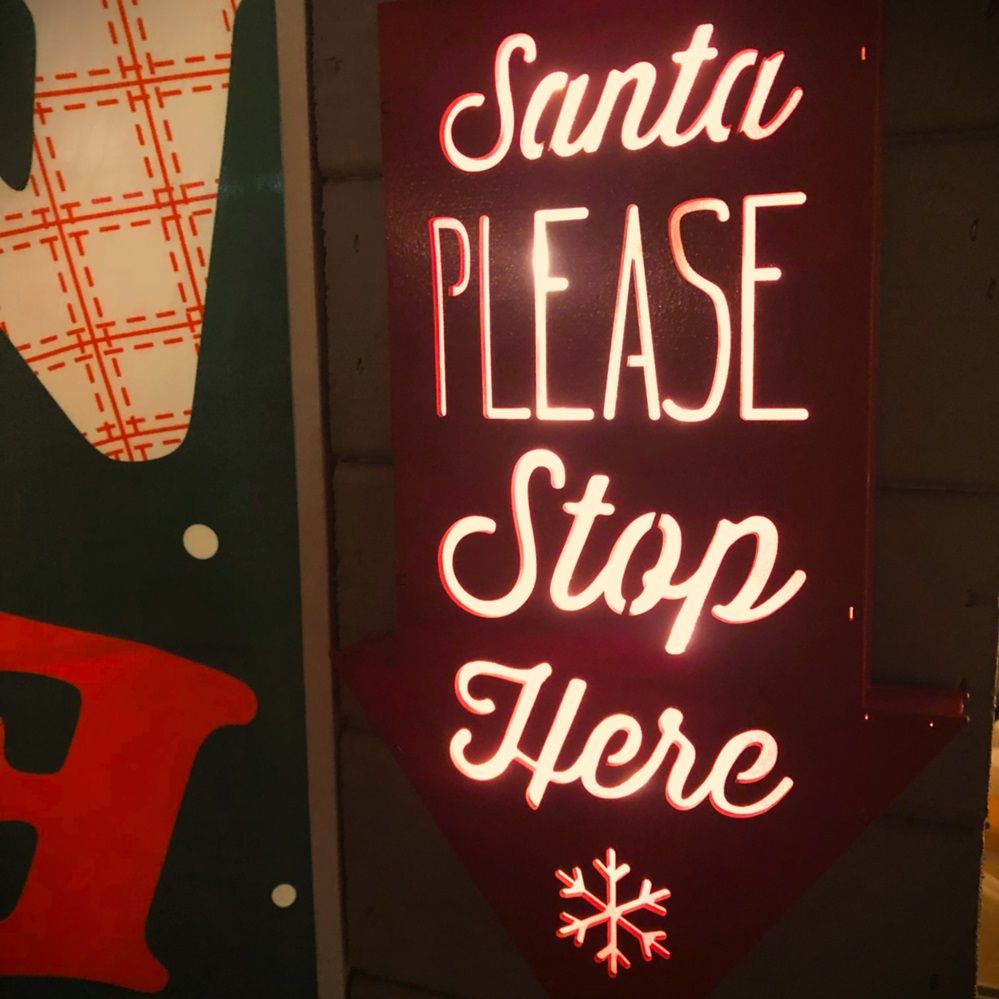 Rotes Pfeilschild mit weißem Text „Santa PLEASE Stop Here“ und einer Schneeflocke unten.
