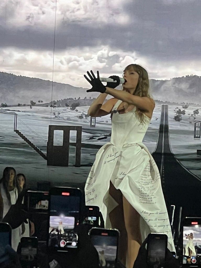Sängerin (Taylor Swift) tritt in einem weißen Outfit auf und singt vor einer schneebedeckten Bergkulisse in ein Mikrofon.