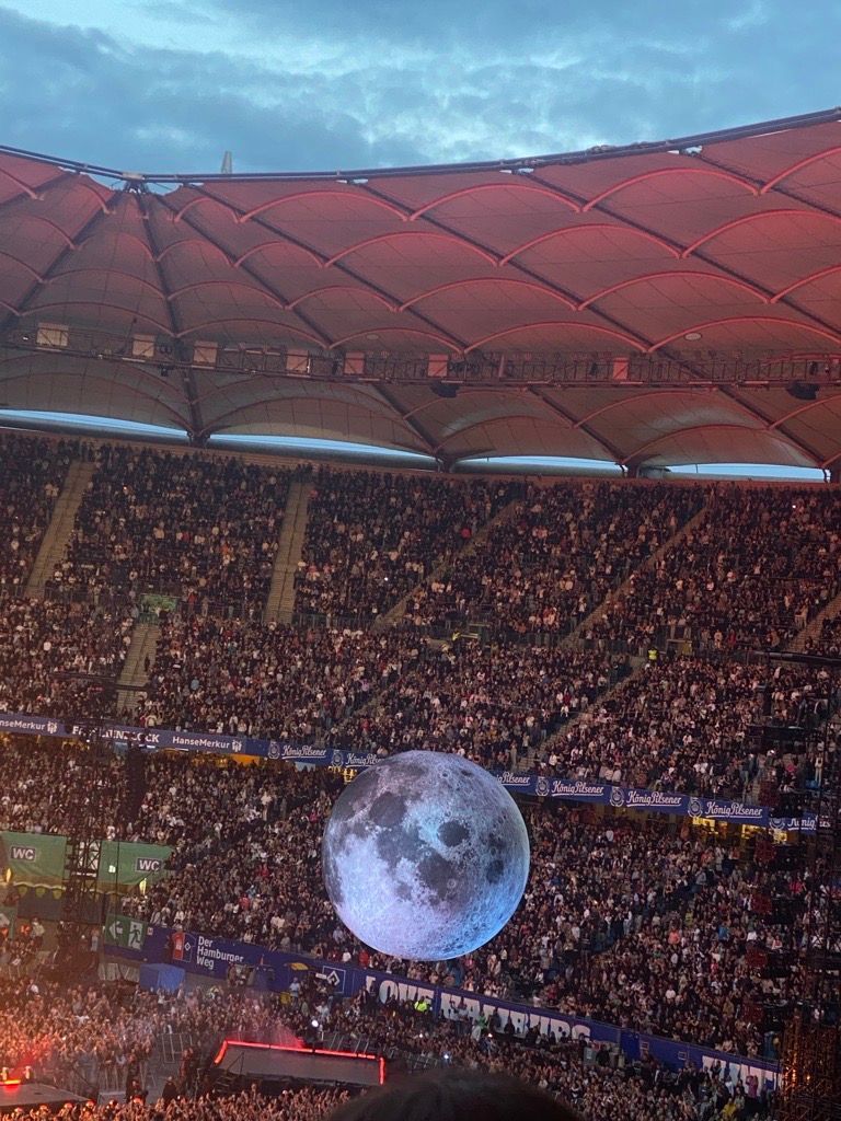 Ein vollbesetztes Stadion bei Nacht, über der Menge unter einem roten Baldachin eine leuchtende, mondähnliche Kugel prangt.