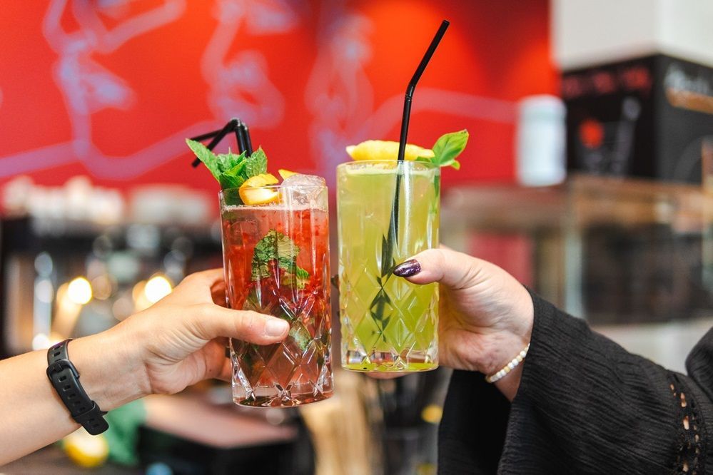 Zwei Hände stoßen mit farbenfrohen Cocktails an, die mit Minze garniert sind, in einer lebhaften Bar.