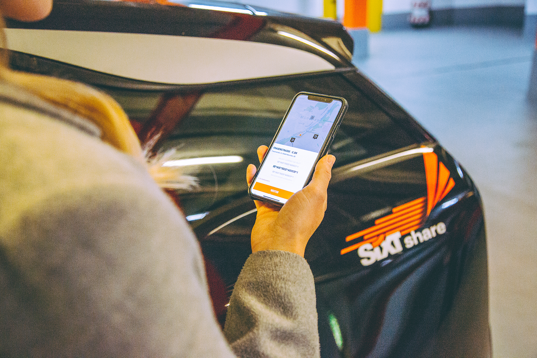 Person using a phone to unlock a Sixt share car in a parking garage; orange Sixt logo visible on car.